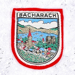 May include: A rectangular embroidered patch with a bright red border featuring the text BACHARACH at the top. The design depicts a colourful landscape with a blue river, green hills, a tall church spire, and various small buildings in a town.