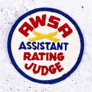 May include: Embroidered patch with a white background, blue border, and red and blue text. The text reads "AWSF ASSISTANT RATING JUDGE" with yellow crossed skis in the center. A vintage-style patch.