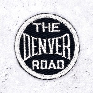 May include: A circular embroidered patch with a black background and a white border. The text THE DENVER ROAD is stitched in white thread across the center, with the word DENVER appearing in a larger, arched font between THE and ROAD.