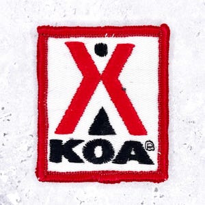 May include: A rectangular embroidered patch with a white background and a red border. The design features a large red X shape above a black triangle and the black text KOA with a small registered trademark symbol in the bottom right corner.