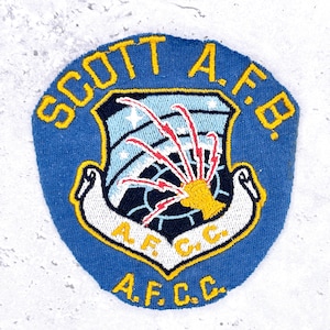 Op de afbeelding: Een blauwe geborduurde patch met de tekst SCOTT A.F.B. en A.F.C.C. in het geel. Het midden toont een schild met een lichtblauwe lucht, witte sterren en een gele hand die rode bliksemschichten uitreikt. Een witte banner bevat de tekst A.F.C.C.