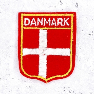 May include: Embroidered patch featuring the Danish flag design. The patch has a red background with a white cross, a yellow border, and the word "DANMARK" in white letters.