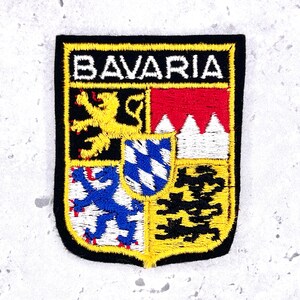 Könnte beinhalten: Ein rechteckiger gestickter Aufnäher mit dem Wort BAVARIA in weißem Garn am oberen Rand. Das Wappen ist in vier Quadranten unterteilt, die einen gelben Löwen, rote und weiße Zacken, einen blauen Löwen und schwarze Formen zeigen, mit einem blau-weißen Rautenwappen in der Mitte.