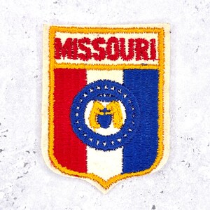 Pode incluir: Patch bordado com a palavra "MISSOURI" em vermelho, acima de um desenho de escudo. O escudo é dividido verticalmente em seções vermelhas, brancas e azuis, com um círculo azul contendo um emblema amarelo. O patch tem uma borda amarela.
