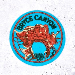 May include: A round patch with a light blue background and a red border. The patch features the text "BRYCE CANYON" in green letters. The center of the patch has an embroidered image of a red and brown rock formation.