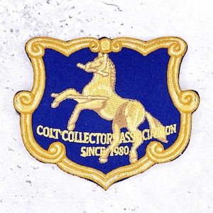 May include: A shield-shaped embroidered patch featuring a blue background with a gold border. In the centre, a gold horse stands on its hind legs. The text COLT COLLECTOR ASSOCIATION SINCE 1980 is embroidered in gold across the lower section.
