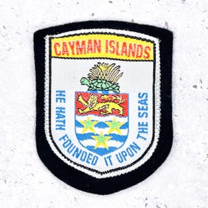 Peut inclure: Un écusson en forme de bouclier avec une bordure noire, affichant les mots "CAYMAN ISLANDS" en orange. L'écusson présente un blason avec une tortue, un lion et des étoiles, et le texte "HE HATH FOUNDED IT UPON THE SEAS."