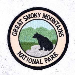 Puede incluir: Un parche bordado circular que muestra un oso negro sentado sobre una roca gris frente a montañas verdes y un cielo azul claro. El borde es negro y el anillo exterior de color crema contiene el texto GREAT SMOKY MOUNTAINS NATIONAL PARK.