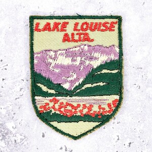Könnte beinhalten: Gestickter Aufnäher mit der Aufschrift "LAKE LOUISE ALTA." Der Aufnäher zeigt eine Berglandschaft mit grünem Rand, einem violetten Gebirgszug, grünen Bäumen und einem roten See. Der Aufnäher ist schildförmig.