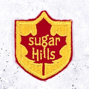 May include: A shield-shaped embroidered patch featuring a red maple leaf silhouette on a red background with a thick yellow border. The text "sugar Hills" is embroidered in yellow across the center of the maple leaf design.