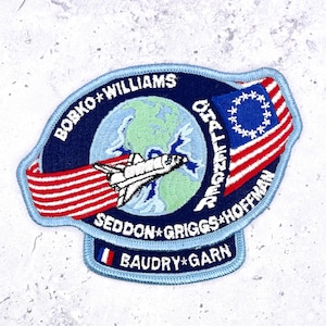 Könnte beinhalten: Ein kreisförmiger gestickter Aufnäher mit hellblauem Rand, der ein Space Shuttle über einem Globus zeigt. Der Text lautet BOBKO WILLIAMS CHALLENGER SEDDON GRIGGS HOFFMAN BAUDRY GARN. Er enthält ein rot-weiß gestreiftes Band und eine blaue Flagge mit Sternen.