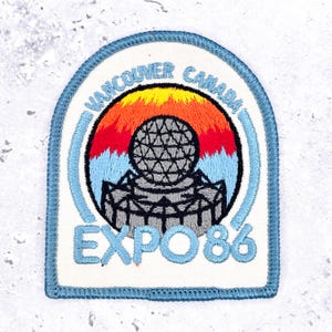 May include: An embroidered patch with a blue border featuring a circular design of a geodesic dome structure against a sunset background of yellow, orange, and red. The text reads VANCOUVER CANADA and EXPO 86 in light blue lettering on a white background.