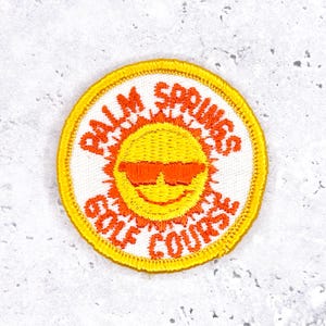 May include: Embroidered patch with a yellow border and a white center. The patch features a smiling sun wearing orange sunglasses, with the words "PALM SPRINGS GOLF COURSE" in orange lettering.