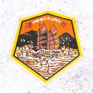 May include: A pentagon-shaped patch featuring a Hong Kong design. The patch has a yellow border and a central image of a traditional Chinese junk sailing on water, with the word "HONGKONG" at the top.