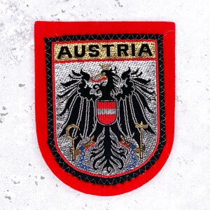 Könnte beinhalten: Ein rechteckiger, roter Stickaufnäher mit abgerundeten unteren Ecken, der das Wort AUSTRIA in schwarzer Schrift auf goldenem Grund zeigt. In der Mitte befindet sich ein schwarzer Adler mit rot-weißem Schild, der Hammer und Sichel hält.