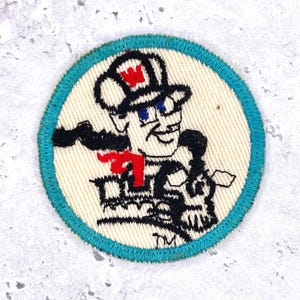 May include: Round embroidered patch featuring a cartoon train conductor with a top hat and a red tie. The patch has a cream-colored background with a teal border and black and red details. The train conductor is smiling.