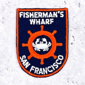 Peut inclure: Écusson brodé bleu marine avec le texte "FISHERMAN'S WHARF SAN FRANCISCO". Le patch a une bordure orange et un motif central de roue de navire orange. Un crabe blanc est au centre de la roue.