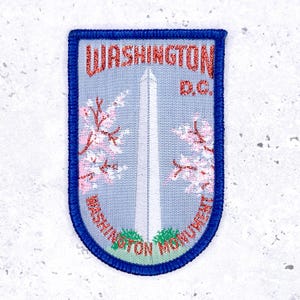 May include: Embroidered patch featuring the Washington Monument and cherry blossoms. The patch has a blue border and includes the text "WASHINGTON D.C." and "WASHINGTON MONUMENT". The background is light blue.