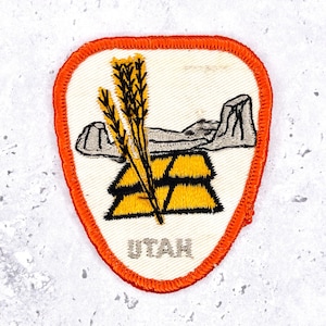 May include: A rectangular patch with an orange border and a white interior. The patch features a stylized depiction of wheat, a rock formation, and stacked gold bars. The word "UTAH" is printed in gray at the bottom of the patch.
