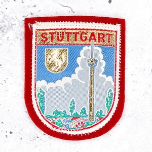 May include: A shield-shaped embroidered patch with a red border. The center features a blue sky with white clouds, a gold shield with a white horse, and a tall tower. The word STUTTGART is embroidered in red on a gold background at the top.