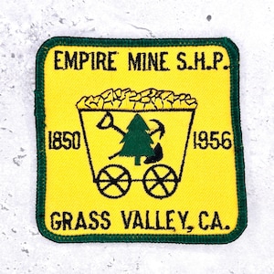 May include: A square embroidered patch with a yellow background and a dark green border. It features a black outline of a mining cart filled with rocks, a green pine tree, a shovel, and a pickaxe. Text reads EMPIRE MINE S.H.P., 1850, 1956, GRASS VALLEY, CA.