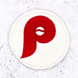 Könnte beinhalten: Ein kreisförmiger bestickter Aufnäher mit cremefarbenem Hintergrund und einer genähten Umrandung. In der Mitte des Aufnähers befindet sich ein großer, kräftiger, roter Buchstabe P mit einem stilisierten, geschwungenen Design.