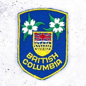 May include: A shield-shaped embroidered patch featuring a blue background with a yellow border. It displays two white dogwood flowers, a crest with a red, white, and blue Union Jack, a yellow sun, and the text BRITISH COLUMBIA in yellow embroidery.