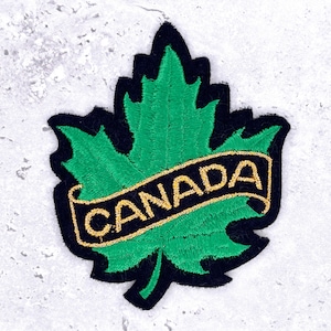May include: Embroidered patch featuring a green maple leaf with a black border. A gold banner across the leaf reads "CANADA". This patch is designed for application on clothing or accessories, symbolizing Canadian identity.