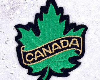 May include: Embroidered patch featuring a green maple leaf with a black border. A gold banner across the leaf reads "CANADA". This patch is designed for application on clothing or accessories, symbolizing Canadian identity.