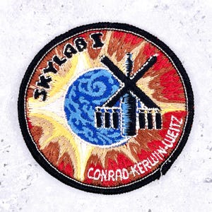 May include: A circular embroidered patch featuring a blue Earth graphic centered against a yellow and red sunburst background. A black silhouette of the Skylab space station is overlaid. The text SKYLAB 1 and CONRAD-KERWIN-WEITZ is stitched.