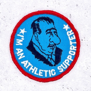 Pode incluir: Um patch redondo com um centro azul apresentando uma ilustração de desenho animado do rosto de um homem. O texto "I'M AN ATHLETIC SUPPORTER" está em branco ao redor do centro azul, com uma borda vermelha.