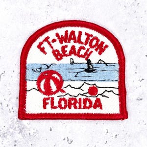 Può includere: Toppa ricamata con bordo rosso e le parole "Ft. Walton Beach Florida" in rosso. La toppa presenta una scena di spiaggia con un oceano blu, un pallone da spiaggia rosso e sagome di persone in acqua.