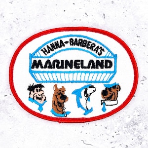 May include: An embroidered patch featuring the text "HANNA-BARBERA'S MARINELAND" in blue and black, with a red border. The patch includes cartoon characters, including a brown dog, a blue dolphin, and a bear.