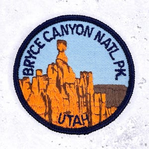 Puede incluir: Parche bordado con el Parque Nacional Bryce Canyon en Utah. El parche circular tiene un borde azul con el nombre del parque. El centro muestra formaciones rocosas naranjas sobre un fondo azul claro, con "UTAH" en naranja.