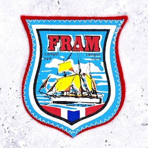 May include: A shield-shaped embroidered patch with a red border. The centre features a sailing ship with yellow sails on a light blue background. The text reads FRAM, Lengst syd, and Lengst nord. A small red, white, and blue flag is at the bottom.