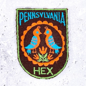 Può includere: Toppa ricamata con sfondo marrone, con due uccelli arancioni con dettagli blu che si fronteggiano. La toppa ha un bordo verde e le parole "PENNSYLVANIA" e "HEX" in blu e verde.