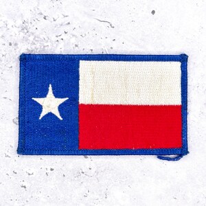Può includere: Una toppa ricamata rettangolare con il design della bandiera dello stato del Texas. La toppa presenta una striscia verticale blu sul lato sinistro con una singola stella bianca a cinque punte e strisce orizzontali bianche e rosse con bordo blu.