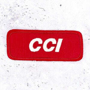 May include: A rectangular red embroidered patch with rounded corners featuring the white text CCI stitched in the centre. The patch has a visible stitched border around the entire perimeter, creating a clean and defined edge for the fabric design.
