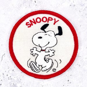 May include: A circular embroidered patch featuring a white background with a red border. The design depicts a cartoon dog with black ears and a red collar, dancing. The text SNOOPY is embroidered in red letters above the character's head.