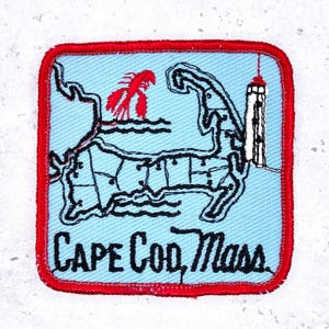 Op de afbeelding: Een vierkante patch met een lichtblauwe achtergrond en een rode rand. De patch toont een kaart van Cape Cod, Massachusetts, met een rode kreeft en een vuurtoren. De tekst "CAPE COD, Mass" is in het zwart gedrukt.