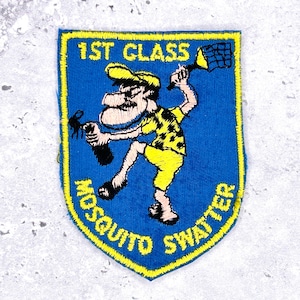 Op de afbeelding: Een blauw en geel geborduurde patch met de tekst "1ST CLASS MOSQUITO SWATTER". De patch toont een stripfiguur in gele kleding die muggen wegvangt met een net. De patch heeft een gele rand.