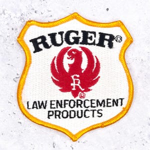 Pode incluir: Um emblema bordado em forma de escudo com fundo branco e borda amarela. Apresenta o texto preto RUGER, um logótipo de fénix vermelho e o texto preto LAW ENFORCEMENT PRODUCTS. O design está centrado no emblema de tecido.