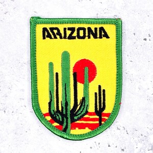 May include: Embroidered patch with the word "ARIZONA" above a desert scene. The design includes green cacti, a red sun, and red and yellow ground. The patch has a green border. Ideal for jackets or bags.