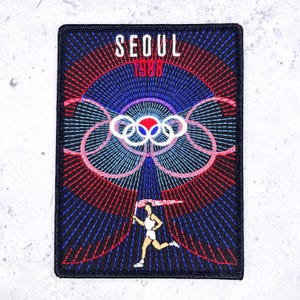 May include: A rectangular embroidered patch featuring the text SEOUL 1988 in white and red. The design includes the Olympic rings in white, blue, and red, set against a dark blue background with radiating red and blue lines and a runner.