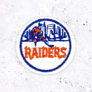 Könnte beinhalten: Ein runder, weißer Aufnäher mit blauem Rand. Der Aufnäher zeigt eine orangefarbene und blaue Grafik eines Hockeyspielers und einer Stadtlandschaft. Das Wort "RAIDERS" ist orangefarben und unten gebogen.