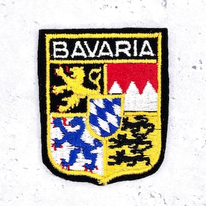 May include: Embroidered patch featuring the word "BAVARIA" above a shield design. The shield has a black border and a yellow outline. The shield contains a yellow lion, a red and white mountain scene, a blue and white diamond pattern, and a blue lion.