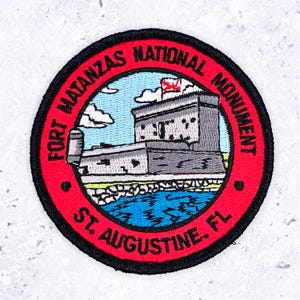 Könnte beinhalten: Ein runder bestickter Aufnäher mit einer grauen Steinfestung und einer roten Flagge vor hellblauem Himmel und dunkelblauem Wasser. Der rote Rand enthält den schwarzen Text FORT MATANZAS NATIONAL MONUMENT und ST. AUGUSTINE, FL.