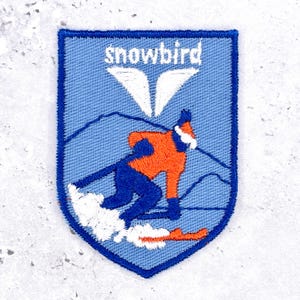 Peut inclure: Un écusson brodé en forme de bouclier sur fond bleu clair. Il représente un skieur avec une veste orange et un pantalon bleu, de la neige blanche projetée et le contour d'une montagne. Le texte "snowbird" est brodé en blanc au-dessus de deux ailes.
