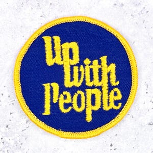 May include: A circular embroidered patch featuring a deep blue background with a bright yellow border. The text Up with People is stitched in yellow thread across the center in a stylized, bold font with a distinct, textured appearance.