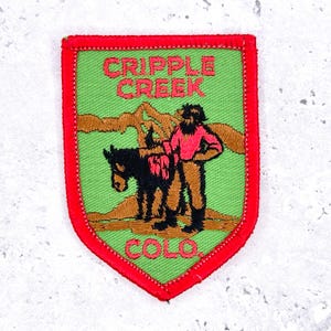May include: Embroidered patch with a red border, featuring the text "CRIPPLE CREEK COLO." in red. The patch depicts a man in a pink shirt and brown pants standing next to a black horse, with a green background and brown mountains.